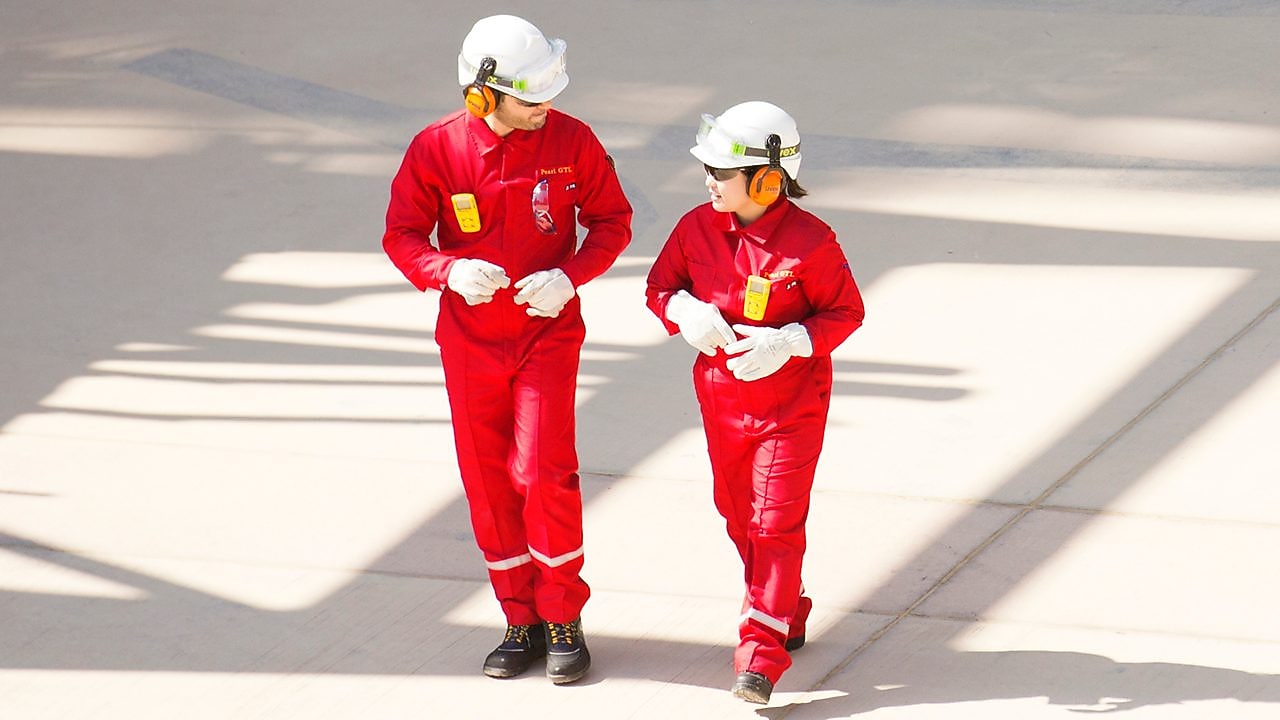 Trabajador y trabajadora con overoles rojos, cascos blancos y protección auditiva caminando