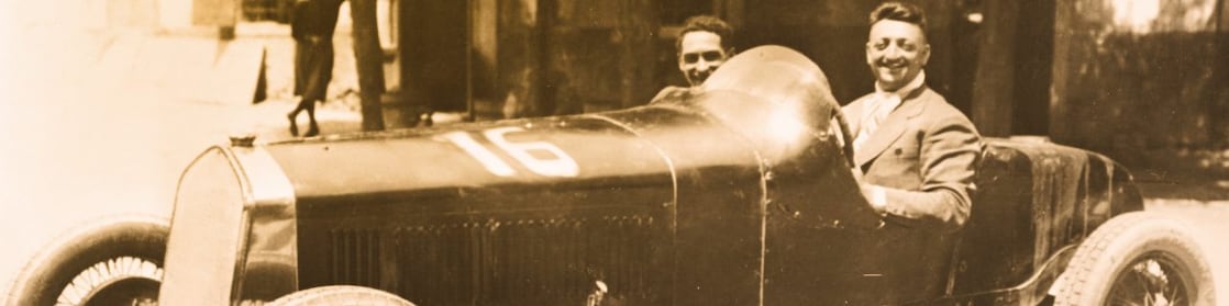 Fotografía en blanco y negro de Enzo Ferrari sentado en uno de sus primeros autos Ferrari