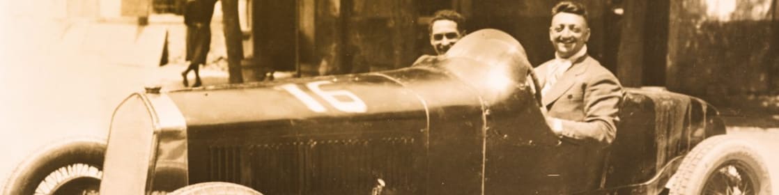 Fotografía en blanco y negro de Enzo Ferrari sentado en uno de sus primeros autos Ferrari