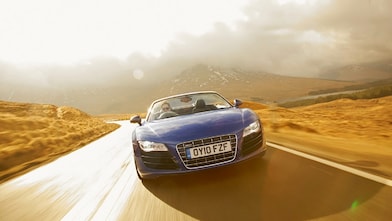 Auto azul descapotable, de cara, circulando por la campiña rodeado de paisajes montañosos