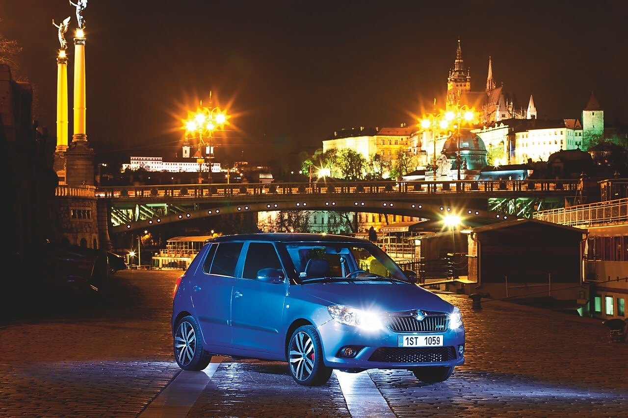 Auto azul estacionado en una calle de Praga con un puente y el castillo al fondo, de noche