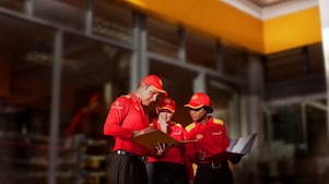 Three employees (one man and two women) in Shell uniform — red shirt and cap — examine a folder the man is holding. One of the women also carries in her hand a briefcase with documents.